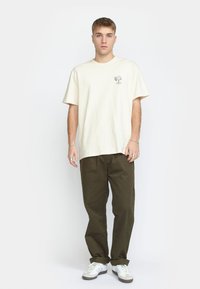 Crème katoenen t-shirt met een kleine zwarte grafiek op de linkerkant van de borst, gecombineerd met donker olijfgroene relaxed-fit broek en witte sneakers.