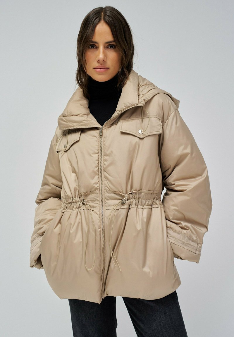 Chaqueta acolchada beige con capucha, cintura ajustable con cordón, bolsillos frontales y cierre de cremallera. Acabado de tela suave con un ligero brillo.