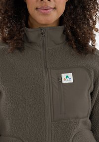 Fleece jacket in olive green with a zip-up design, featuring a chest pocket and a logo patch. Textured fabric with smooth accents.
