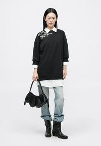 Μαύρη υπερμεγέθη φούτερ μπλούζα με λουλουδάτη κεντητική, layered πάνω από λευκό πουκάμισο με γιακά, συνδυασμένη με ανοιχτό μπλε τζιν και μαύρη τσάντα.