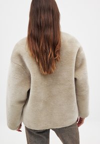 Bežs, pārmērīgi liels mākslīgās kažokādas mētelis ar mīkstu tekstūru un noapaļotiem pleciem. Vienkāršs dizains bez redzamiem aksesuāriem vai rakstiem.