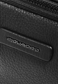 Materiale in pelle nera con finitura testurizzata, caratterizzato da un logo "Piquadro" in rilievo su un elegante elemento rettangolare nero. Dettagli con cerniera ben visibili.