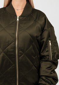 Giacca bomber verde oliva con texture trapuntata, colletto a coste e zip frontale dorata; presenta una tasca con zip sulla manica.