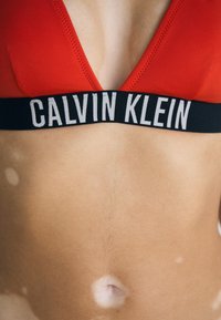 Červený bikiny top s trojuholníkovými pohárikmi, s čiernym pásom označeným "CALVIN KLEIN" v bielych písmenách. Hladká textúra, minimálne švy.
