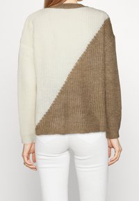 Neulepusero, jossa on vinon muotoinen kerma- ja ruskeakuvio, pyöreä pääntie ja pitkät hihat, teksturoitu kangas ja ribatut hihansuut.