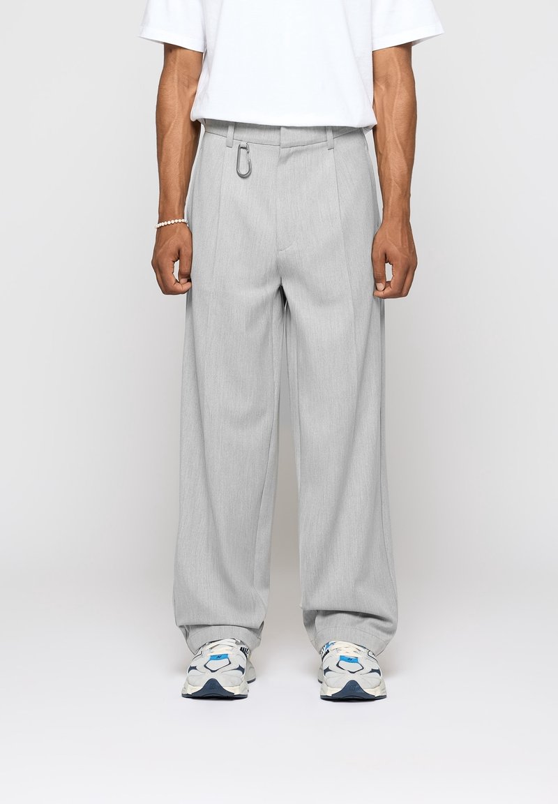 Pantalon gris à jambes larges avec plis et détail anneau métalique à la taille, assorti à des baskets blanches avec des accents bleus.