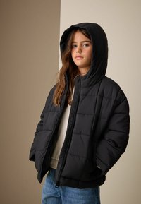 Czarna ocieplana kurtka z kapturem, z pikowanym wzorem i zamkiem błyskawicznym. Noszona na jasnym topie i niebieskich dżinsach.