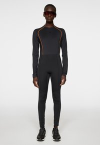 Svart långärmad jumpsuit med orange detaljer, tillverkad av elastiskt material. Figursydd design med sömdetaljer, tillsammans med svarta sportskor.