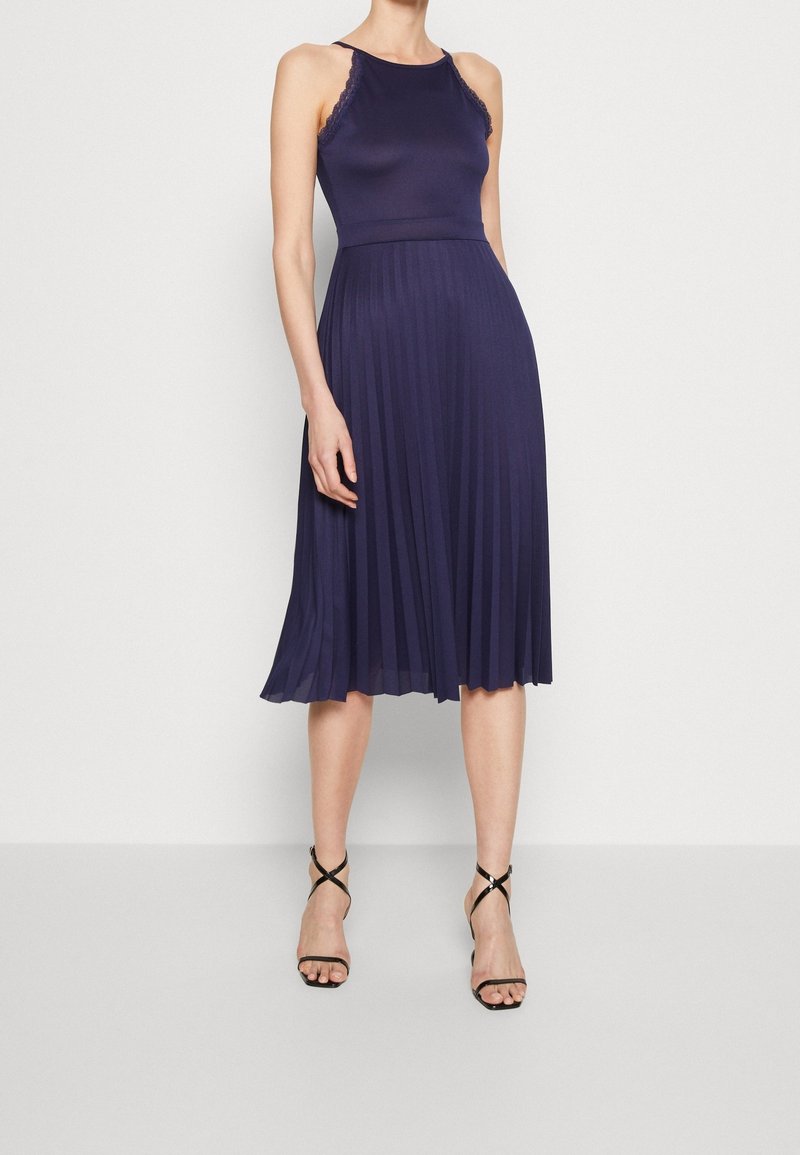 Robe plissée bleu marine avec un corsage ajusté, des détails en dentelle au niveau du col, et une jupe fluide. Des escarpins noirs à lanières complètent la tenue.