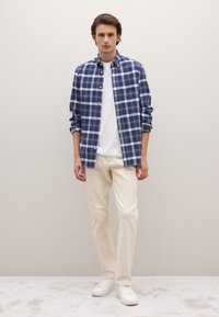 Marineblaue und weiße karierte Hemdjacke über einem weißen T-Shirt, kombiniert mit cremefarbenen Hosen und weißen Turnschuhen, stehend vor einem hellen Hintergrund.