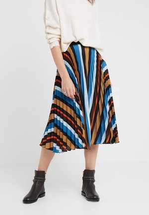 Femme portant une jupe midi plissée avec des rayures diagonales bleues, marron, noires, blanches et orange, des bottines noires et un pull crème.