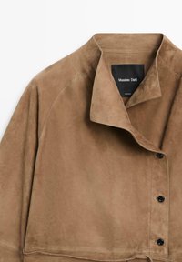 Veste en daim marron clair avec un large col, trois boutons noirs et une étiquette visible "Massimo Dutti" à l'intérieur du col.