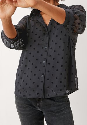 Osoba ubrana w prześwitującą czarną koszulę na guziki w grochy, z podwiniętymi rękawami, w połączeniu z ciemnoszarymi jeansami, z podniesionymi ramionami.