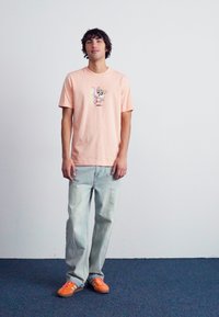 Gaiši rozā kokvilnas T-krekls ar multfilmu grafiku uz krūts. Kombinēts ar gaiši ziliem džinsiem un oranžiem sporta apaviem.