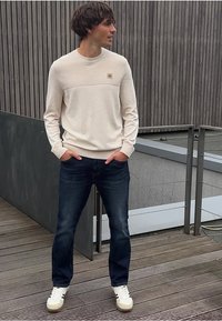 Ljus beige crewneck-tröja med texturerad övre del, kombinerad med mörkblå jeans och vita sneakers. Emblemdetalj på bröstet.