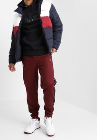 Prošívaná bunda s námořnicky modrými, bílými a červenými panely přes černou mikinu s kapucí; bordó tepláky s bočními pruhy; bílé tenisky.