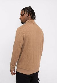 Pull en maille marron avec un col montant, présentant un motif en losanges texturé et un ourlet et des poignets côtelés. La vue arrière montre une coupe ajustée.