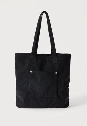 Sort kordfløjls-tote taske med to lange håndtag og en frontlomme sikret med to metalnitter.