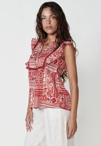Blusa rossa e crema con motivo a fantasia e maniche volan, caratterizzata da design intricati e una vestibilità comoda, abbinata a pantaloni leggeri in lino.
