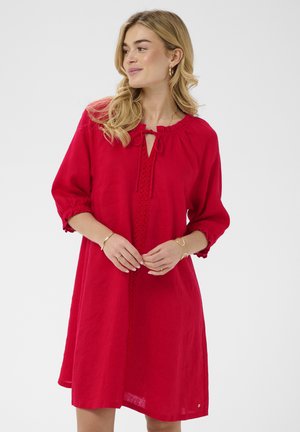 Žena v červenom šatôčkach po kolená s 3/4 rukávmi, čipkovanými detailmi vpredu a viazaním na výstrihu, stojaca na bielom pozadí.