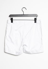 Pantalones cortos de algodón blancos con un frente plano, puños doblados y bolsillos laterales, colgados de un gancho de plástico negro sobre un fondo blanco.