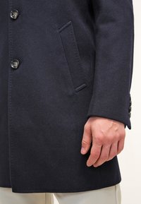 Marinblå ullblandad kappa med knappar, en enda bröstficka och skräddarsydd passform. Tyget har en mjuk textur med minimala sömdetails.