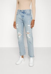 Ljusblå slitna jeans med fransade nederkanter, med flera hål. Matchas med vita sneakers för en avslappnad look.