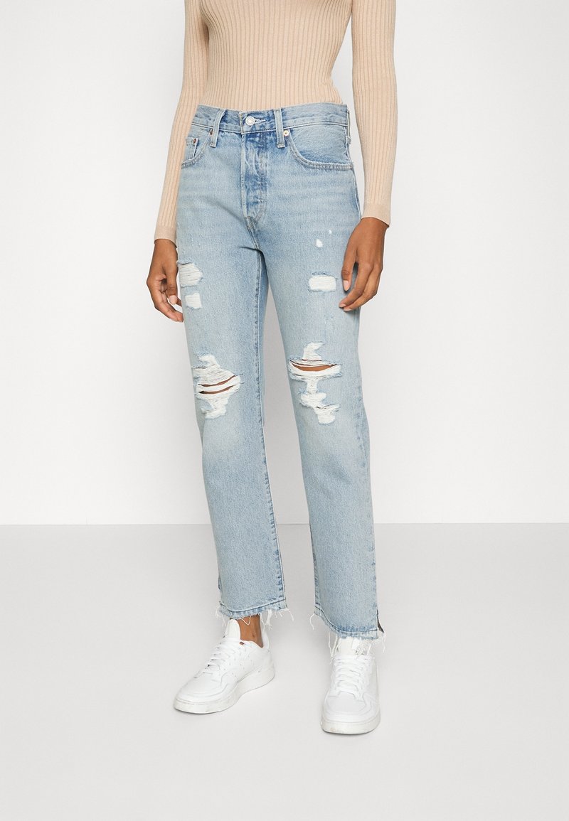 Ljusblå slitna jeans med fransade nederkanter, med flera hål. Matchas med vita sneakers för en avslappnad look.