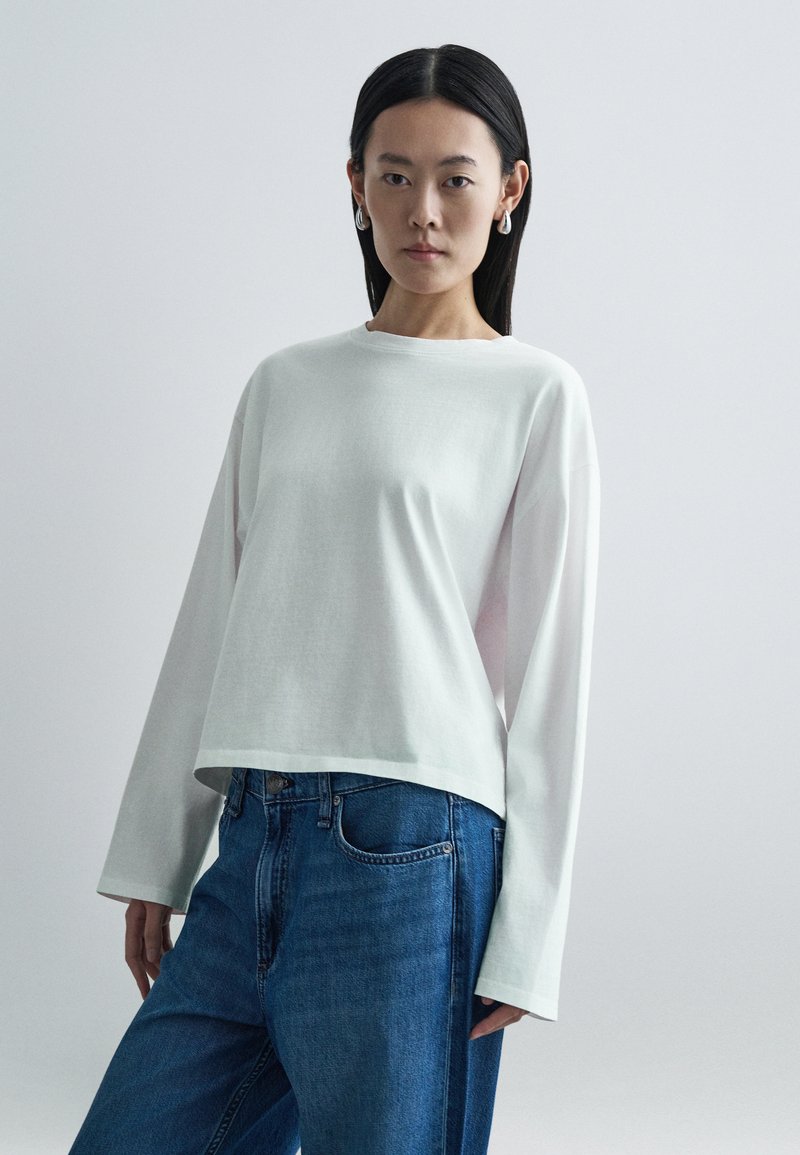 Biała koszulka z długim rękawem o luźnym kroju i okrągłym dekolcie, zestawiona z niebieskimi dżinsami. Koszulka ma gładką fakturę i minimalistyczny design.