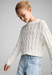 Weißer, kurzer Pullover mit Zopfmuster, langen Ärmeln, geripptem Saum und offener Webstruktur, kombiniert mit hellblauen, hochgeschnittenen Jeans.