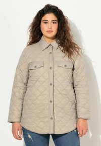 Chaqueta de color beige acolchada con cuello, cierre de botones en la parte delantera y dos bolsillos en el pecho. Presenta una textura suave y un dobladillo redondeado.