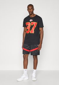 Mand, der står iført en sort Kansas City Chiefs t-shirt med nummer 87, sorte og røde Nike shorts, hvide Nike sokker og hvide sneakers.