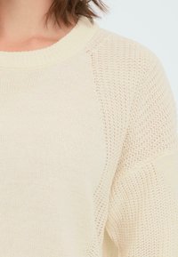 Lysbeige strikkegenser med ribbet rund hals, teksturerte raglanermer og en løs passform. Stoffet virker mykt og pustende.
