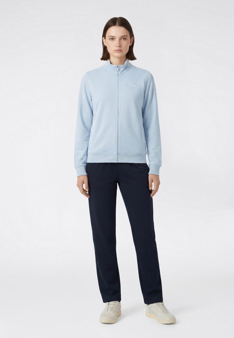 Femme debout portant une veste zippée bleu clair, un pantalon bleu marine et des baskets blanches sur un fond blanc uni.