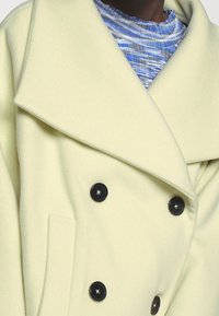 Světlě žlutý dvouřadý kabát s velkými klopami, černými knoflíky a měkkou texturou. Obsahuje skrytý kapsu na boku.