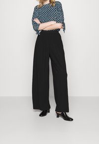 Pantalons larges noirs en tissu texturé, associés à un blouse bleu et blanc à motif, avec des manches nouées. Chaussures à talons noirs.