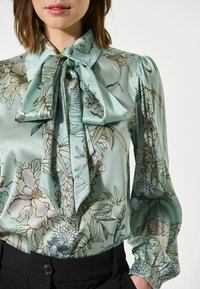 Blusa floreale azzurro chiaro con colletto a fiocco, maniche a sbuffo e polsini elastici; presenta una texture lucida e motivi botanici bordati di marrone.