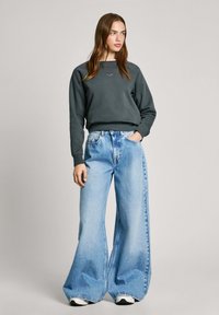 Mörkgrå sweatshirt med rund hals i kombination med ljusblå vida byxor, vilket visar en lös passform och mjuk tygtextur. Vita sneakers fulländar looken.
