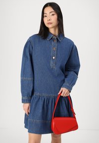 Denimkjole med krage, knappestolpe og samlet skjørt. Modellen holder en elegant rød håndveske. Stoffet er teksturert og blått.
