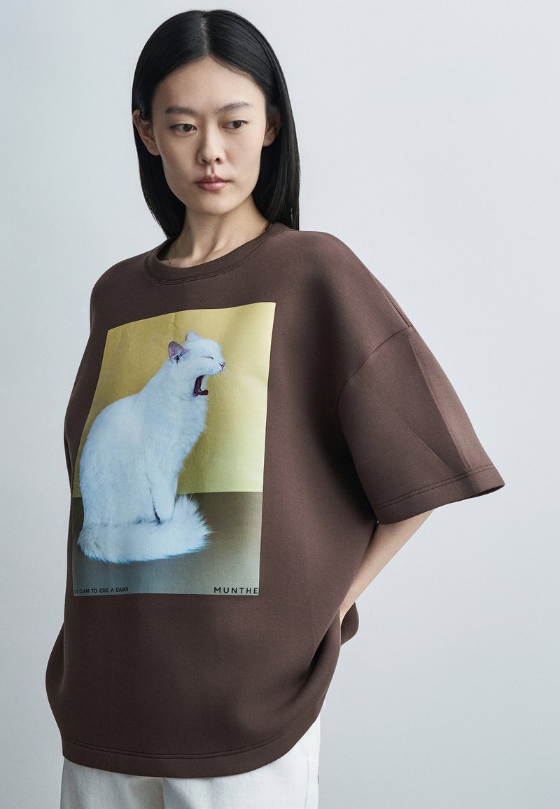 Γυναίκα με ίσια μαύρα μαλλιά φορά φαρδιά καφέ μπλούζα με μεγάλη εικόνα μιας χασμουρητής λευκής γάτας σε κίτρινο φόντο.