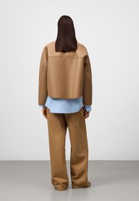Braune kurz geschnittene Jacke mit glatter Textur, über einem hellblauen Hemd getragen. Lockere braune Hose ergänzt das Outfit. Glatter Hintergrund.