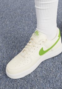 Bijela Nike Air Force 1 tenisica s zelenim naglascima, glatka kožna gornja strana, perforirana prednja strana i gumeni potplat, u paru s bijelim čarapama.
