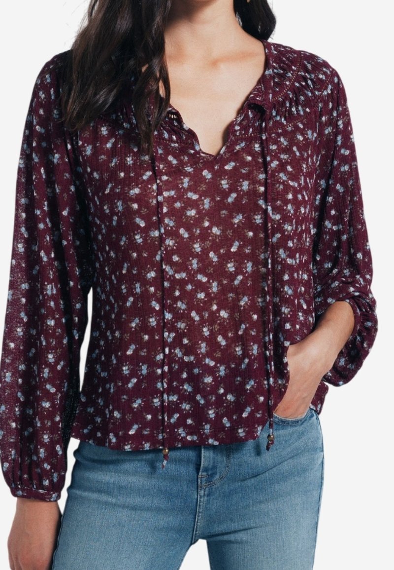 Femme portant un chemisier à manches longues imprimé floral bordeaux avec un col en V et des liens, associé à un jean bleu clair, main dans la poche.