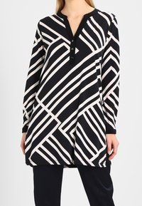 Schwarze Tunika mit weißen diagonal und vertikal gestreiften Mustern, mit einem Henley-Ausschnitt, Knopfleiste und langen Ärmeln. Weicher Stoff mit angenehmer Textur.
