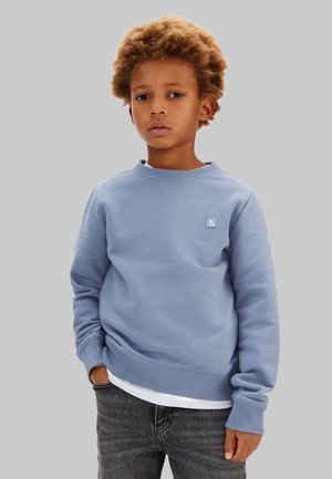 Ung gutt med krøllete hår iført lys blå genser og mørke jeans, står mot en ensfarget grå bakgrunn.