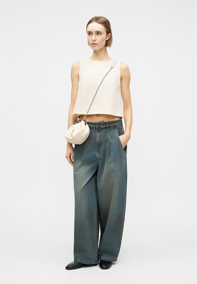 Femme portant un haut court blanc sans manches, un jean large bleu délavé, des chaussures noires et un petit sac bandoulière blanc avec une sangle en chaîne, debout.