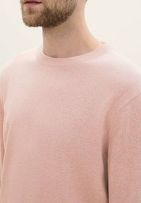 Svetlo roza pleten pulover z okroglim ovratnikom, rebrastimi manšetami in diskretnim logotipom na prsih. Gladka tekstura, prilega se okrog ramen.