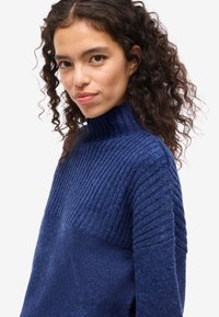 Mėlynas megztinis su aukšta apykakle, turintis raštuotą tekstūrą ir kvadratinę formą. Minkšta medžiaga su kontrastinėmis megztomis dėmėmis ant rankovių.