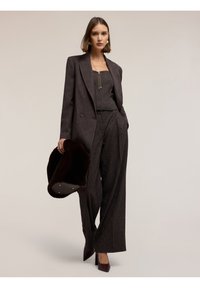 Blazer su misura marrone scuro sopra un pantalone a gamba larga abbinato, abbinato a un top aderente, tacchi alti e una clutch in pelliccia scura. Accessori dorati.