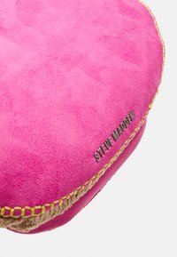 Produit en suède rose avec une surface texturée, présentant un logo métallique "Steve Madden" et un fil jaune contrastant le long du bord.